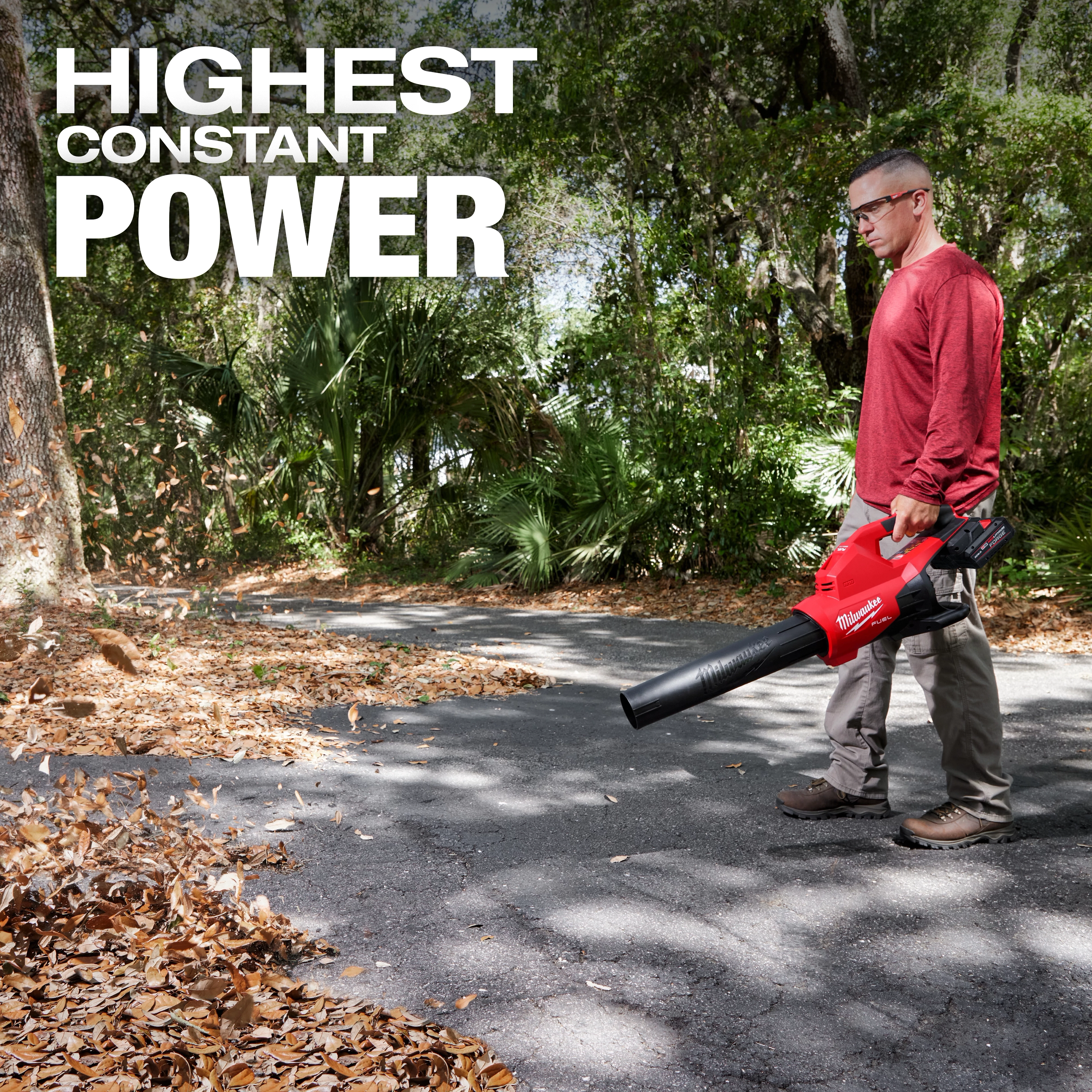A person is using an M18 FUEL™ Dual Battery Blower to clear leaves from a paved path surrounded by trees. The text "HIGHEST CONSTANT POWER" is displayed at the top left of the image.