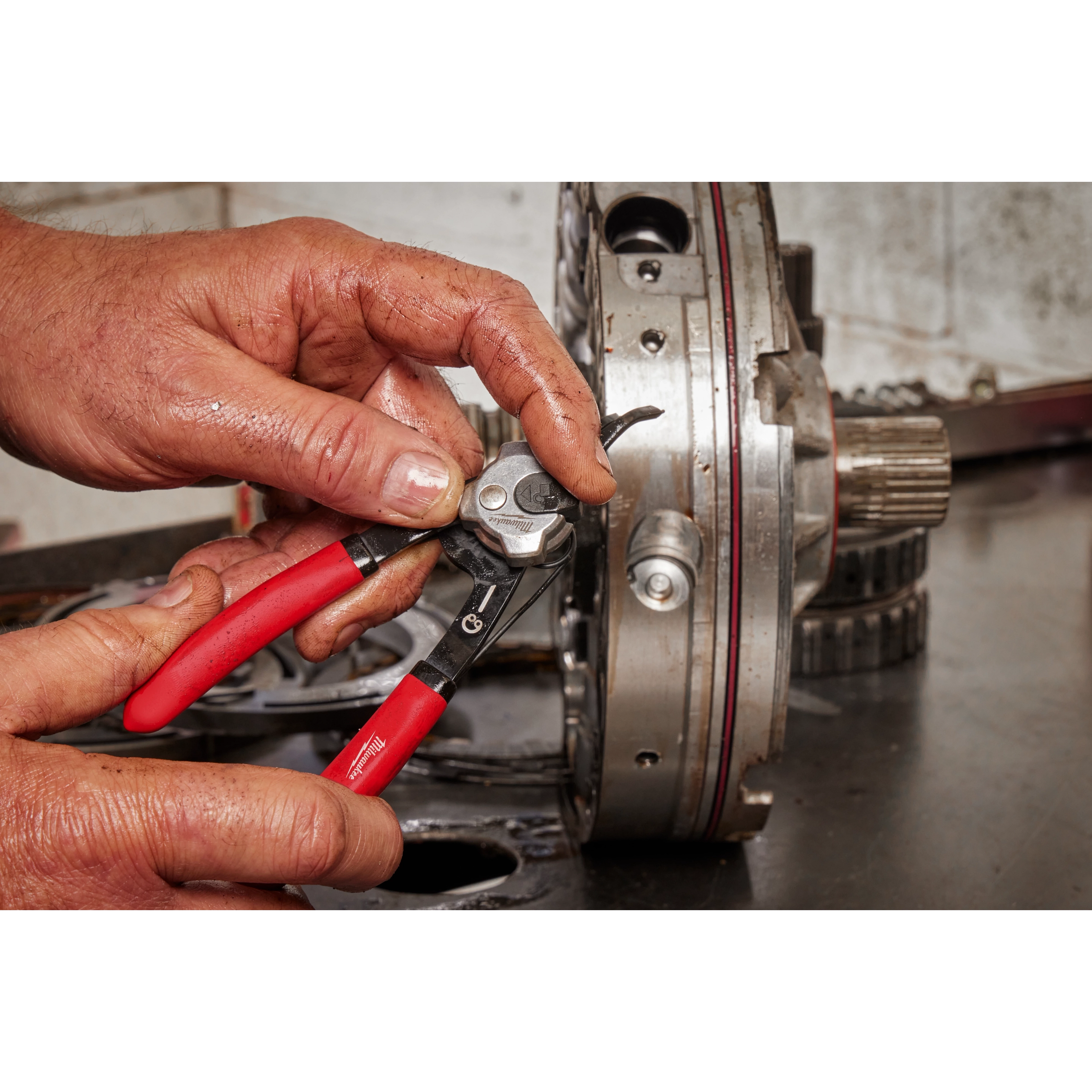 A person uses the Convertible Snap Ring Pliers Set to work on a mechanical component. The pliers have red handles, and the user’s hands are visibly dirty, indicating they are in the middle of a repair task. The mechanical component appears to be part of a larger machine or vehicle.