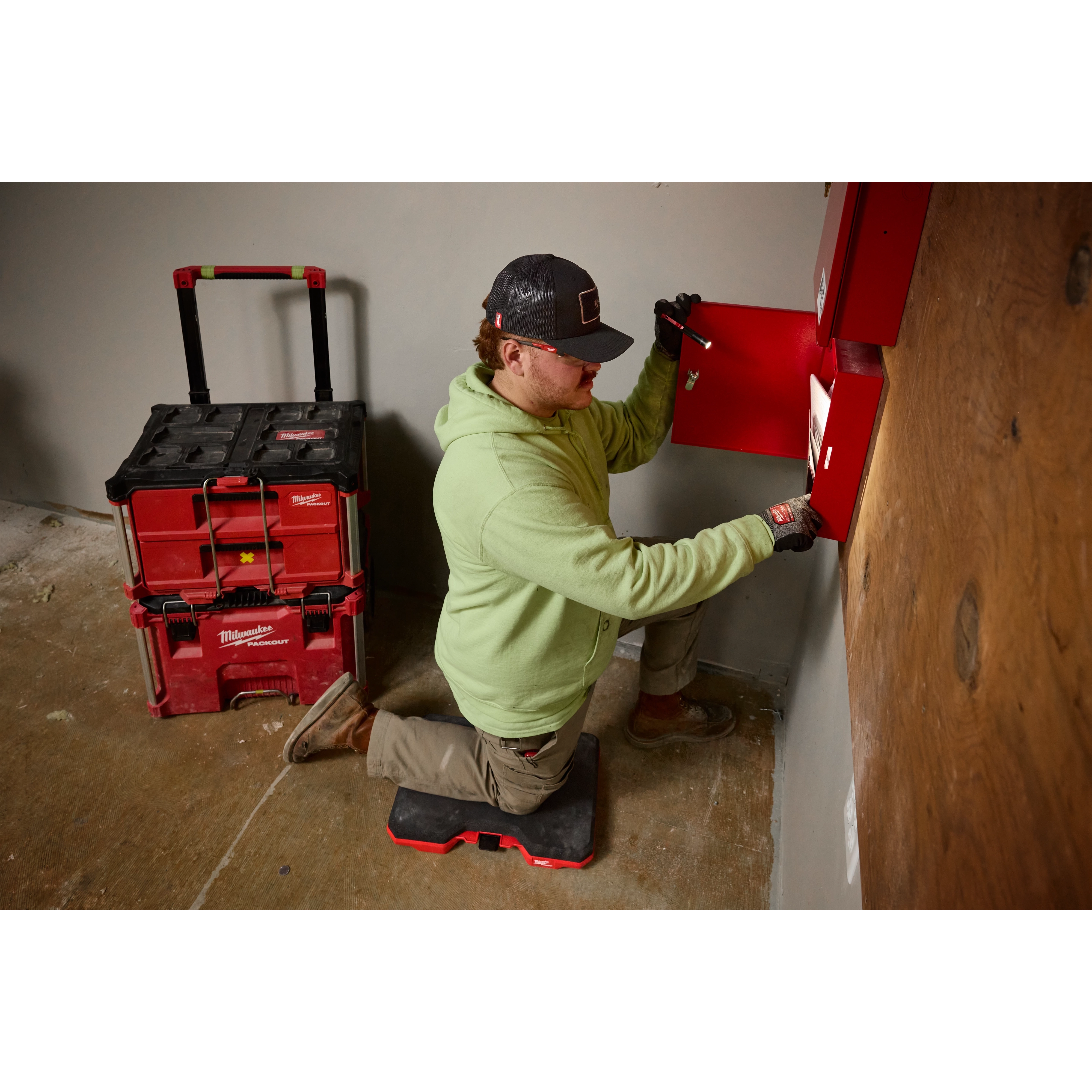 A person wearing a Snapback Bump Cap works on a red wall-mounted unit using a tool. They kneel on a red knee pad beside a stack of red Milwaukee toolboxes on a concrete floor.