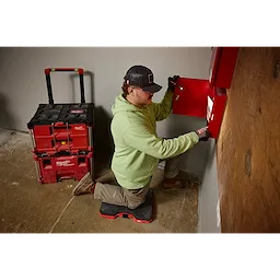 A person wearing a Snapback Bump Cap works on a red wall-mounted unit using a tool. They kneel on a red knee pad beside a stack of red Milwaukee toolboxes on a concrete floor.