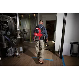 Man using the M18 FUEL™ 3-in-1 Backpack Vacuum to clean a hardwood floor in an industrial setting. The vacuum is red and black, strapped to his back, and connected through a long hose. The room contains large machinery and is partially under construction.