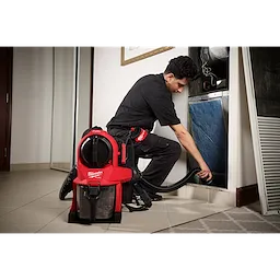 A worker uses a M18 FUEL™ 3-in-1 Backpack Vacuum to clean an HVAC system. The red vacuum is worn on the worker's back and has a black hose attached, ensuring efficient dust removal. The worker wears protective black clothing and kneels beside the HVAC unit.