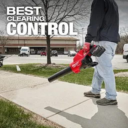 A person uses the M18 FUEL™ Dual Battery Blower to clear a sidewalk. The blower is red and black. The background shows a parking lot with cars, trees, and a building. The text "BEST CLEARING CONTROL" is displayed at the top.