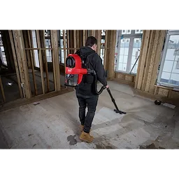 A person uses an M18 FUEL™ 3-in-1 Backpack Vacuum to clean a construction site. The individual wears the red backpack vacuum and operates the hose attachment to clean the floor in a partially framed building.