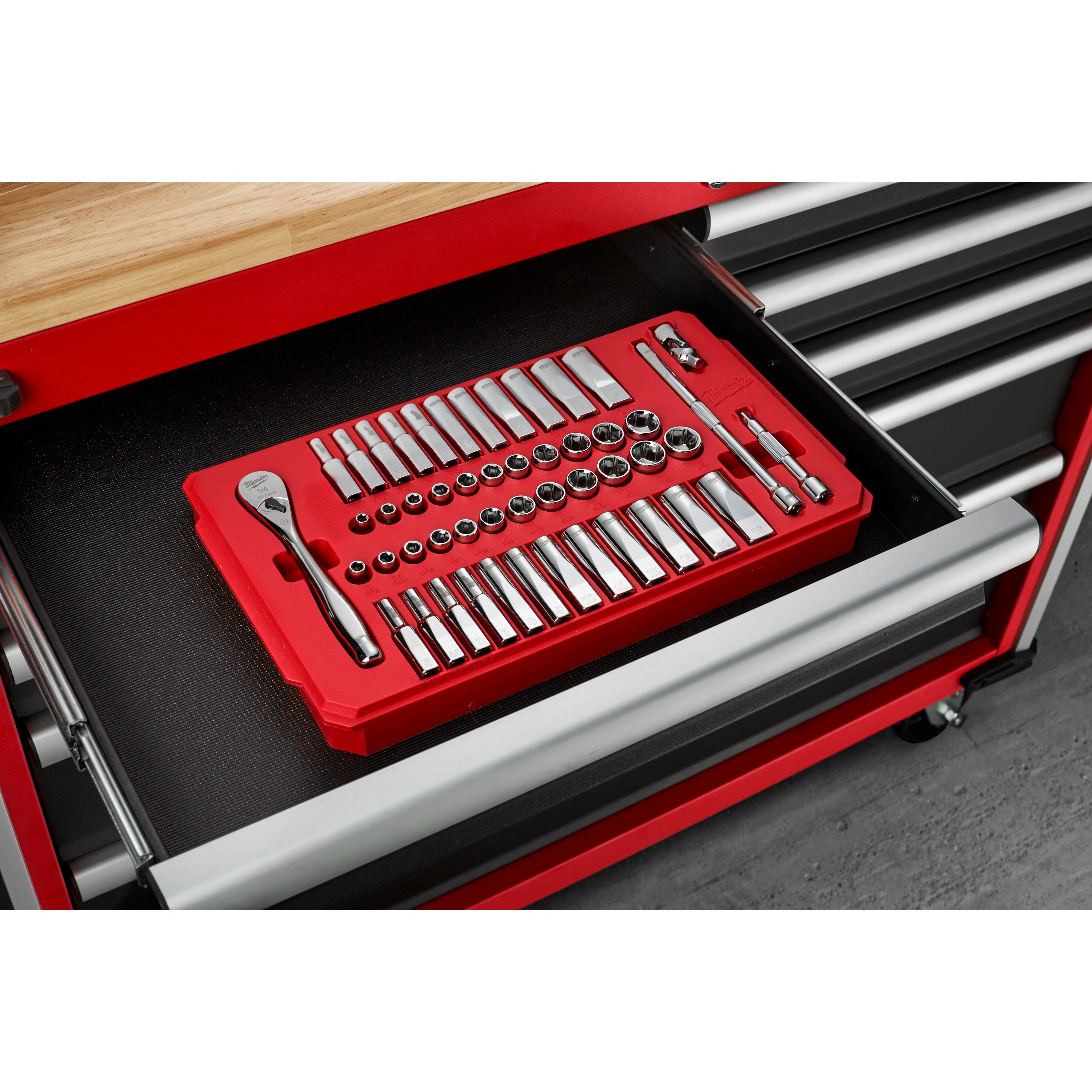 A red tray organizer containing various socket wrenches and accessories is arranged neatly inside an open drawer of a red and black tool chest. The tool chest has multiple drawers and a wooden top surface above the open drawer. The floor beneath is concrete.