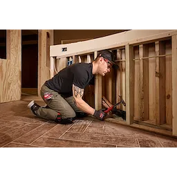 A person wearing a Snapback Bump Cap is kneeling and working on a wooden construction frame. They are using a hammer and gripping a piece of wood in a partially built structure.